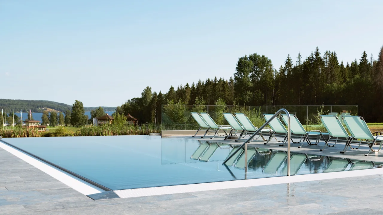 En utomhus infinitypool med solstolar, omgiven av lummiga träd och en lugn sjöutsikt under en klar himmel.