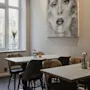 An inviting dining space featuring light wooden tables, patterned chairs, and a large window. A striking black and white portrait adorns the wall.