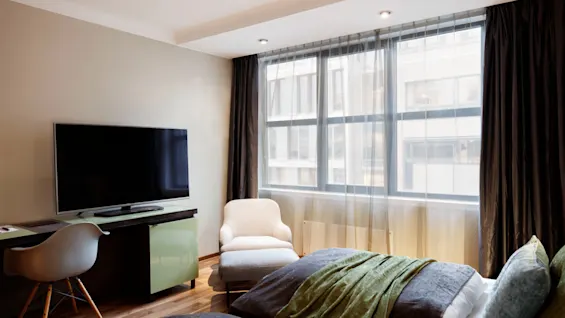 A television on a desk faces a bed in a modern room with large windows. Sheer curtains and darker drapes frame the window, with a white armchair nearby.