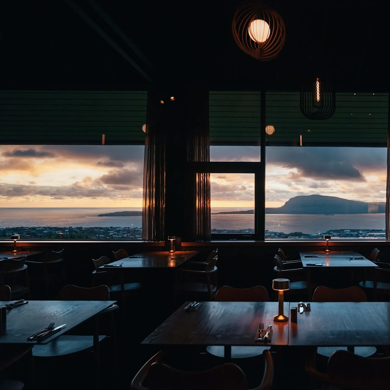 Bordene er dækket til middag i en svagt oplyst restaurant med udsigt over et stort hav og bjerg gennem store vinduer ved solnedgang.