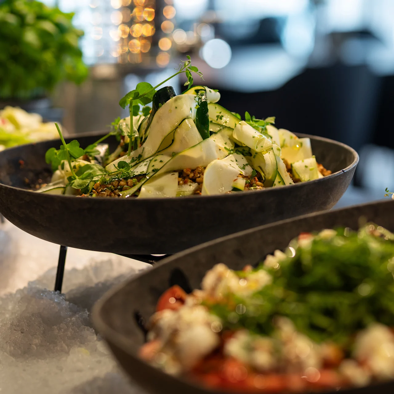 En skål med frisk salat med skåret grøntsager og urter hviler på en seng af is, omgivet af andre retter, indendørs med blød belysning.