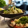 En skål med frisk salat med skåret grøntsager og urter hviler på en seng af is, omgivet af andre retter, indendørs med blød belysning.