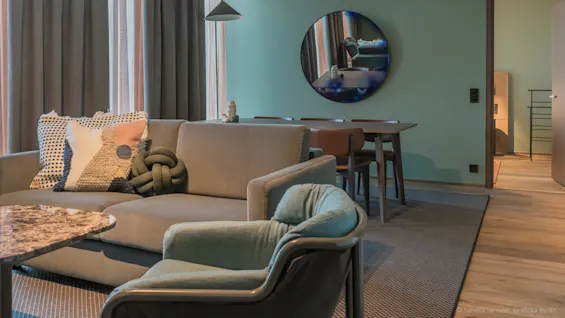 En grå soffa med dekorativa kuddar står i ett modernt vardagsrum med ett marmorbord, stor rund spegel och matplats. Gardiner täcker höga fönster. © Sandra Larsson, Grafiska Byrån