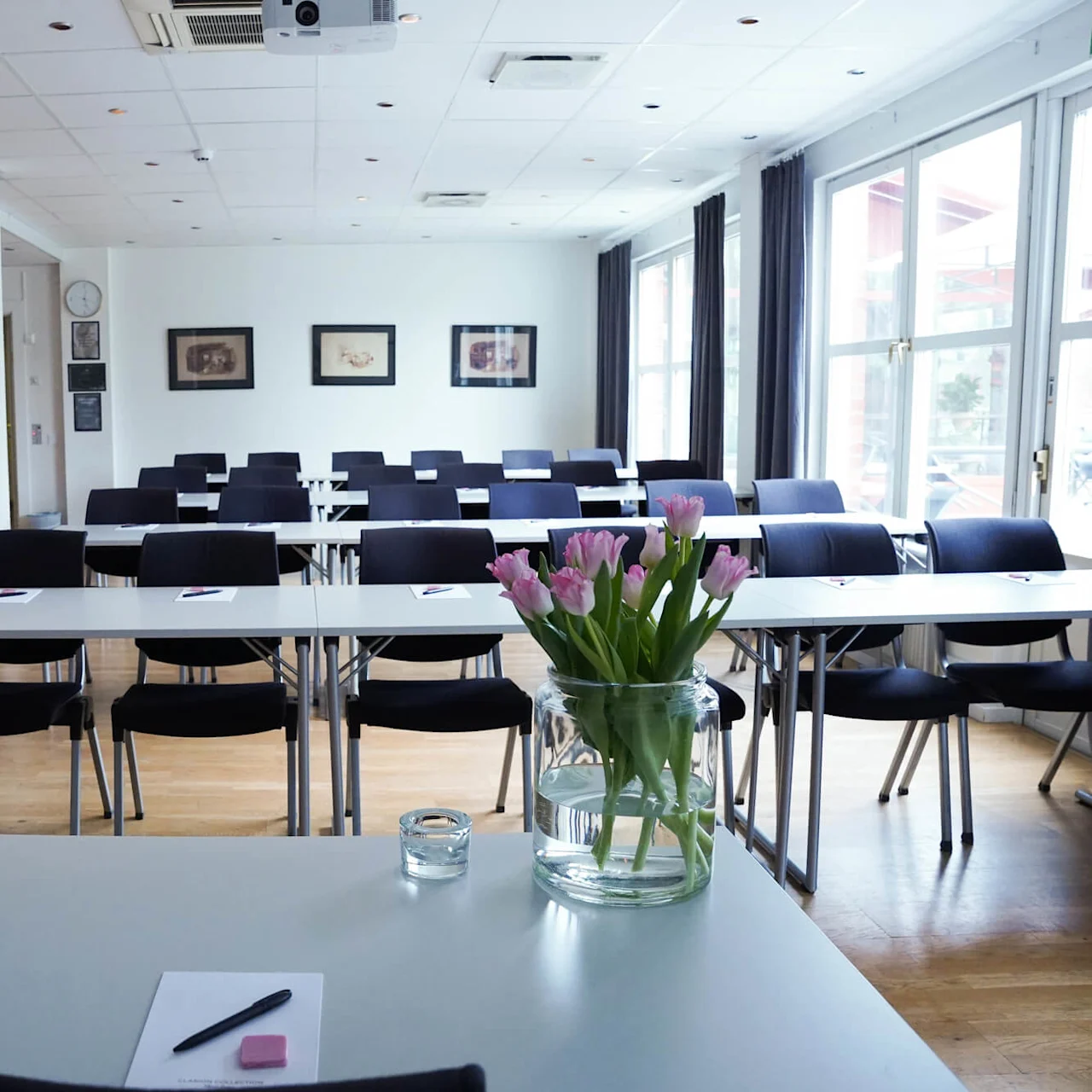Tables and chairs arranged for a meeting or lecture, with a vase of tulips on a table in the foreground, inside a well-lit room.