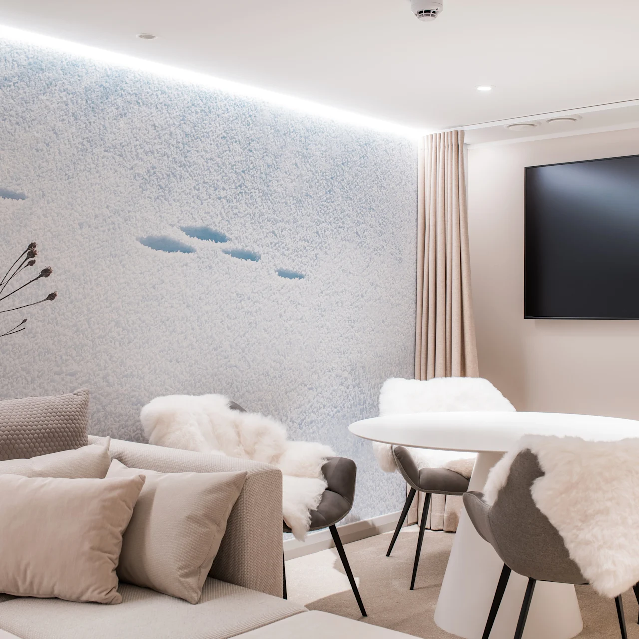 A cozy living space with a round table surrounded by chairs draped with white fur. A large flat-screen TV hangs on the wall, with a textured, snowy mural.