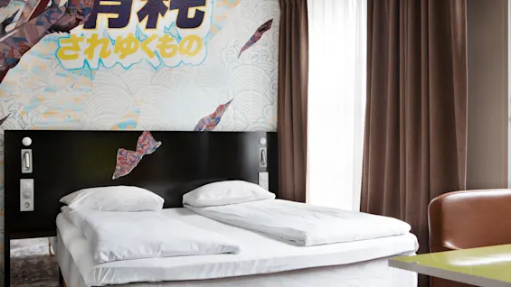 A neatly-made bed with white linens stands against a mural with abstract shapes and Japanese text. Brown curtains and a green table are nearby.
