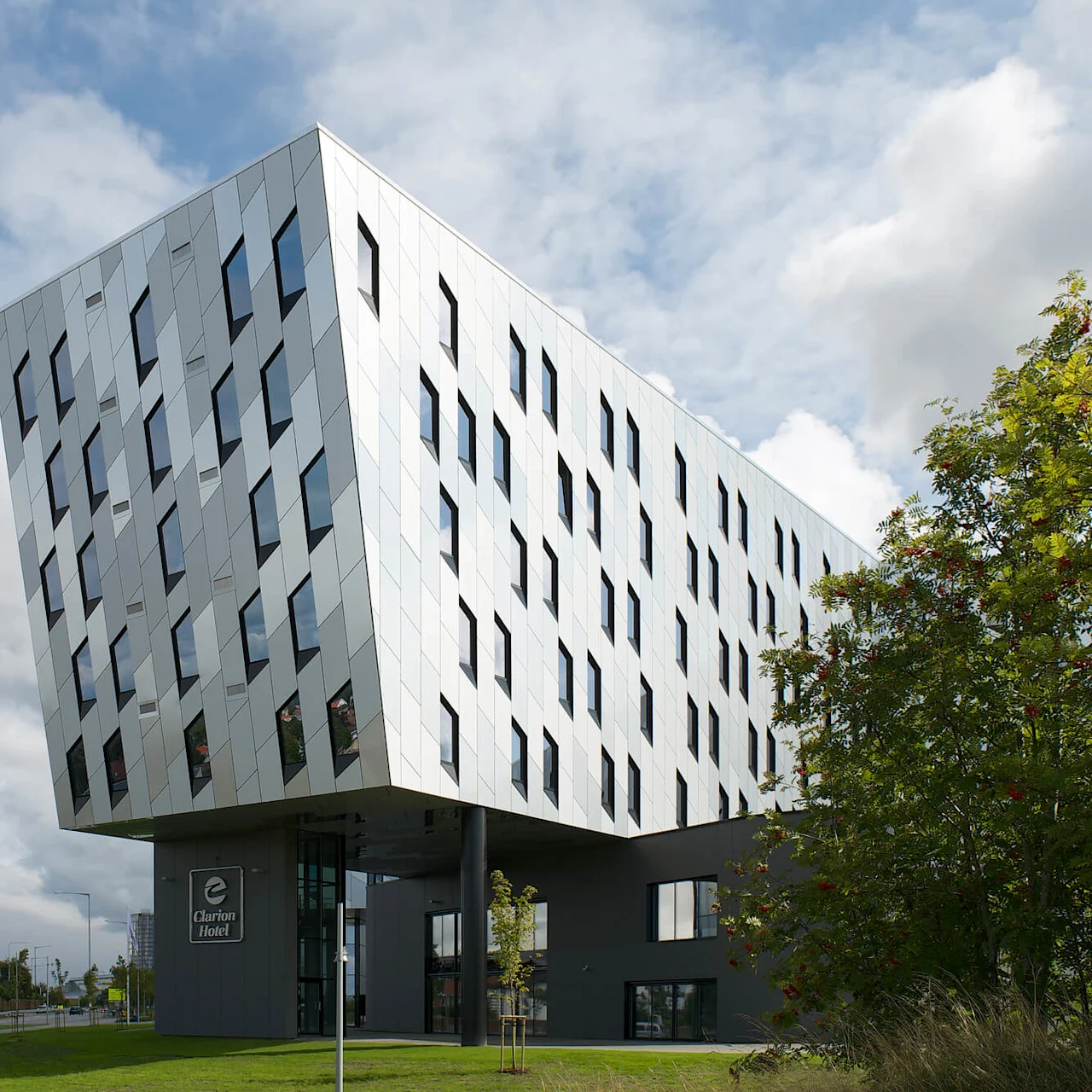En modern, vinklad byggnad lutar över sin bas som stöds av pelare. Fasaden har många fönster. "Clarion Hotel" syns på bottenvåningen. Omgiven av grönt gräs och träd.