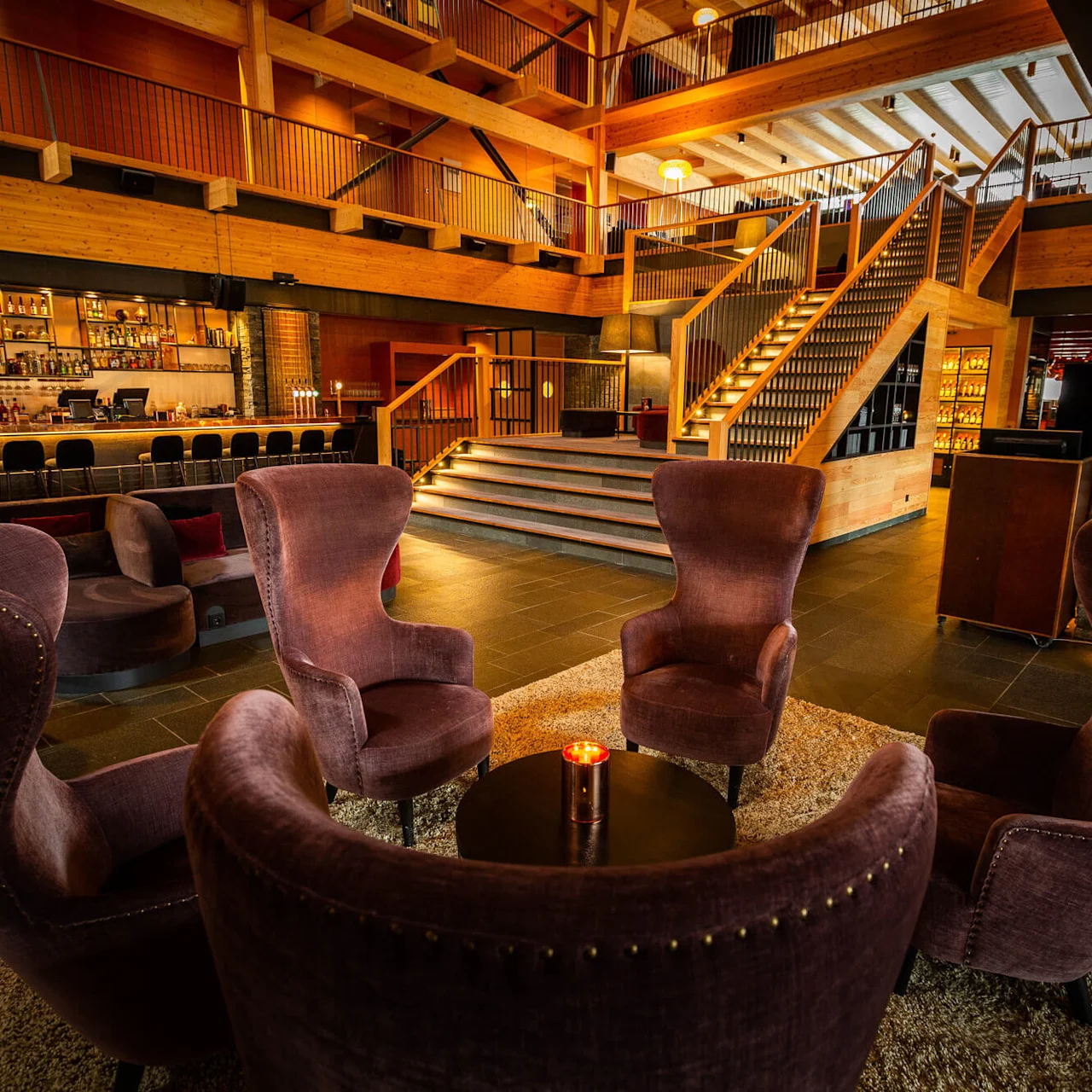 Four plush armchairs encircle a round table with a candle, set on a rug. Behind, a wooden staircase leads upward, with a stocked bar in a warmly lit lounge.