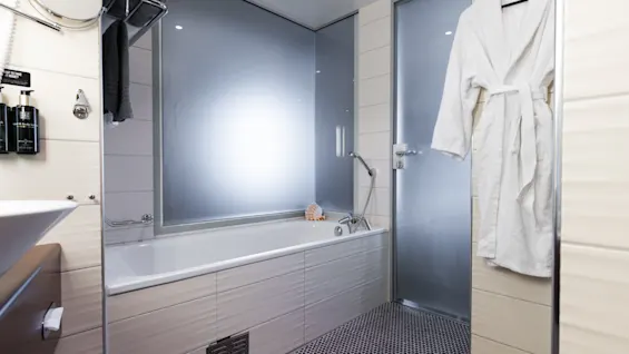 Bathtub with handheld showerhead sits in sleek, modern bathroom; frosted glass window and door separate spaces. Robe hangs nearby, complementing bright, minimalist decor.