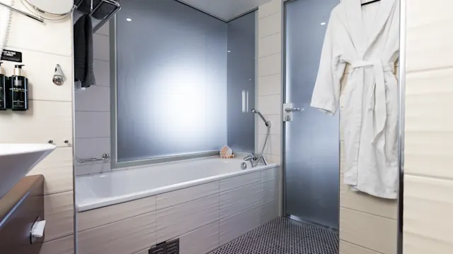 Bathtub with handheld showerhead sits in sleek, modern bathroom; frosted glass window and door separate spaces. Robe hangs nearby, complementing bright, minimalist decor.