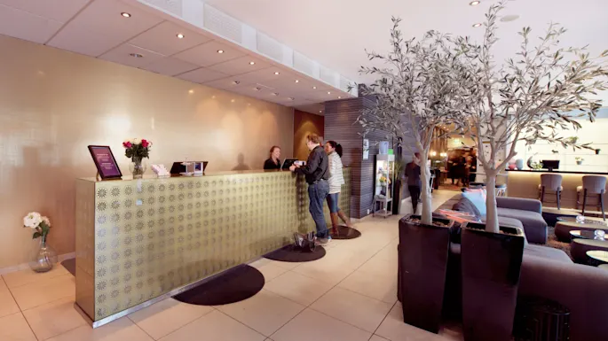 Two guests are checking in at a modern hotel reception desk, with staff assisting them.