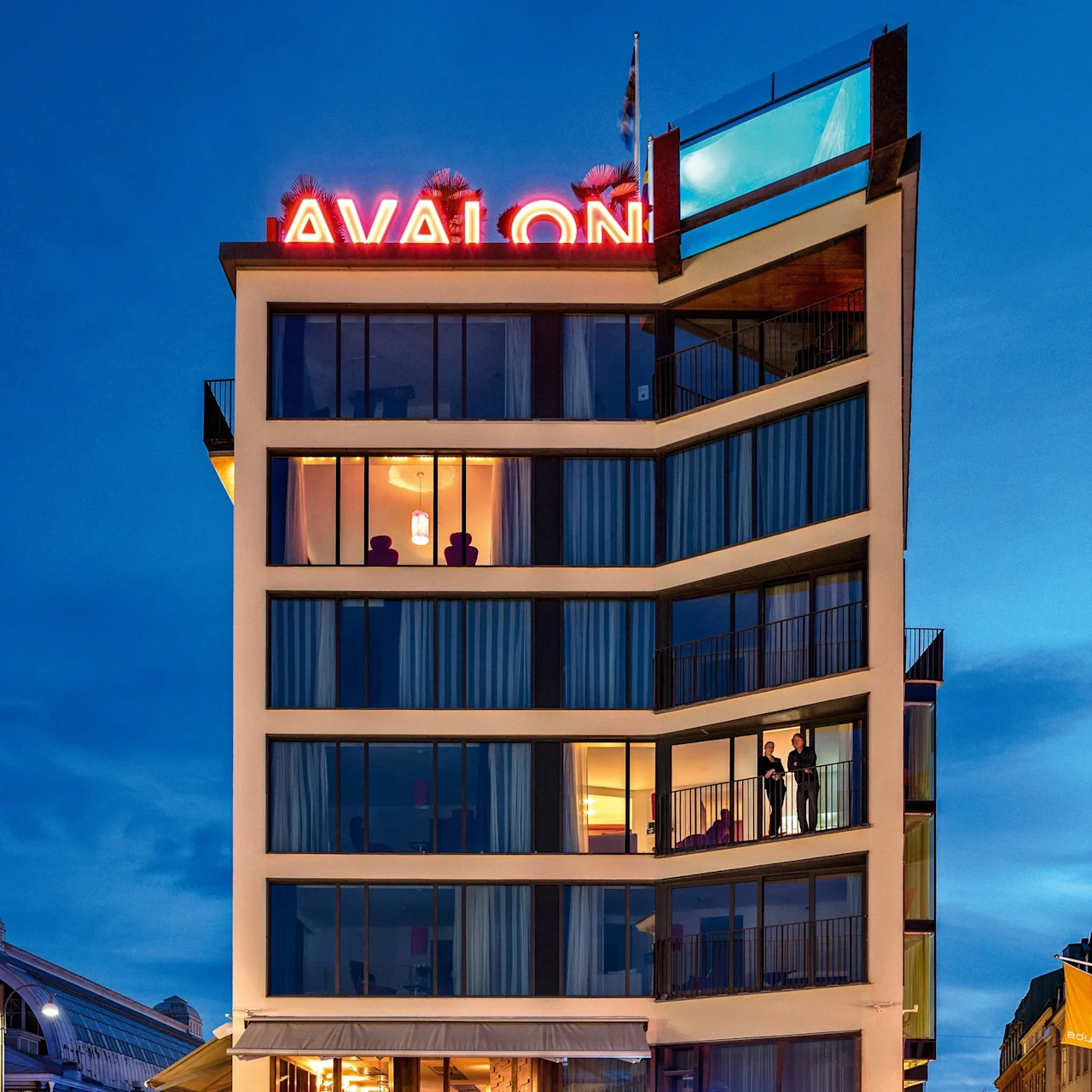 A modern multi-story building with large windows displays "AVALON" in red neon. Two people stand on a balcony. Below, a lit restaurant bar sits in a quiet street.