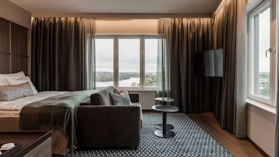 En seng med hvide puder og et grønt tæppe står ved siden af en sofa og et rundt bord i et moderne rum med store vinduer, med udsigt over et naturskønt landskab.