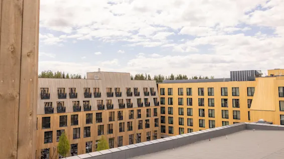 Modernt byggnad med många fönster mot en molnig himmel, sedd från en intilliggande byggnad med synlig träbalk.