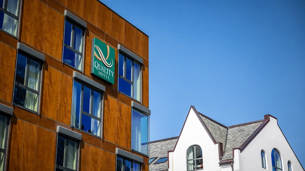Brun bygningfasade viser et skilt med teksten "QUALITY HOTEL" på grønn bakgrunn; ved siden av står en hvit bygning med gråt tak under en klar blå himmel.