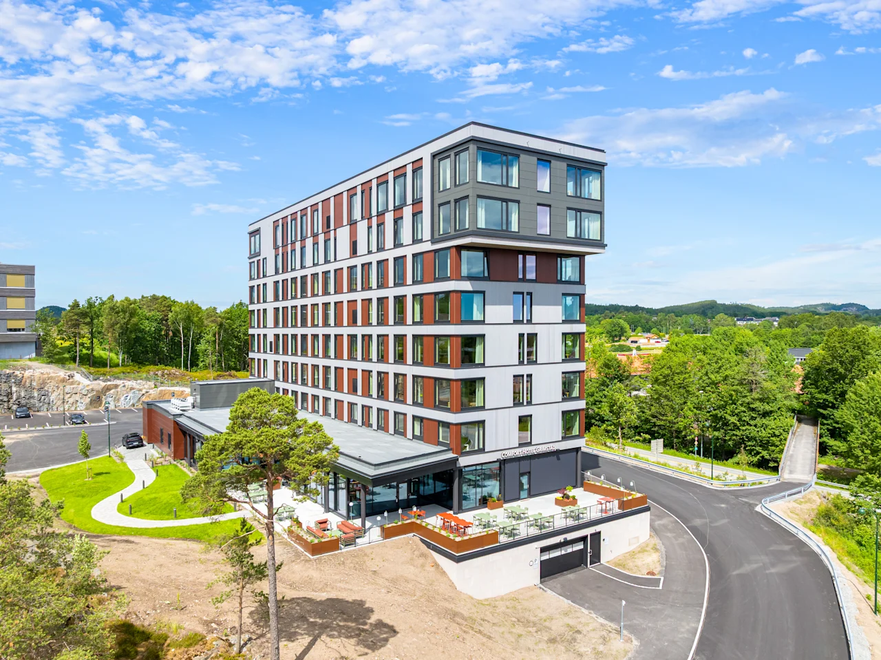 A modern hotel building with a unique design, surrounded by lush green trees and a winding road under a blue sky.