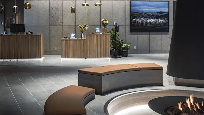 A circular fireplace emits warmth in a modern lobby with curved seating. Wooden reception desks hold flowers and tech devices under pendant lights. A landscape photo decorates the concrete wall.
