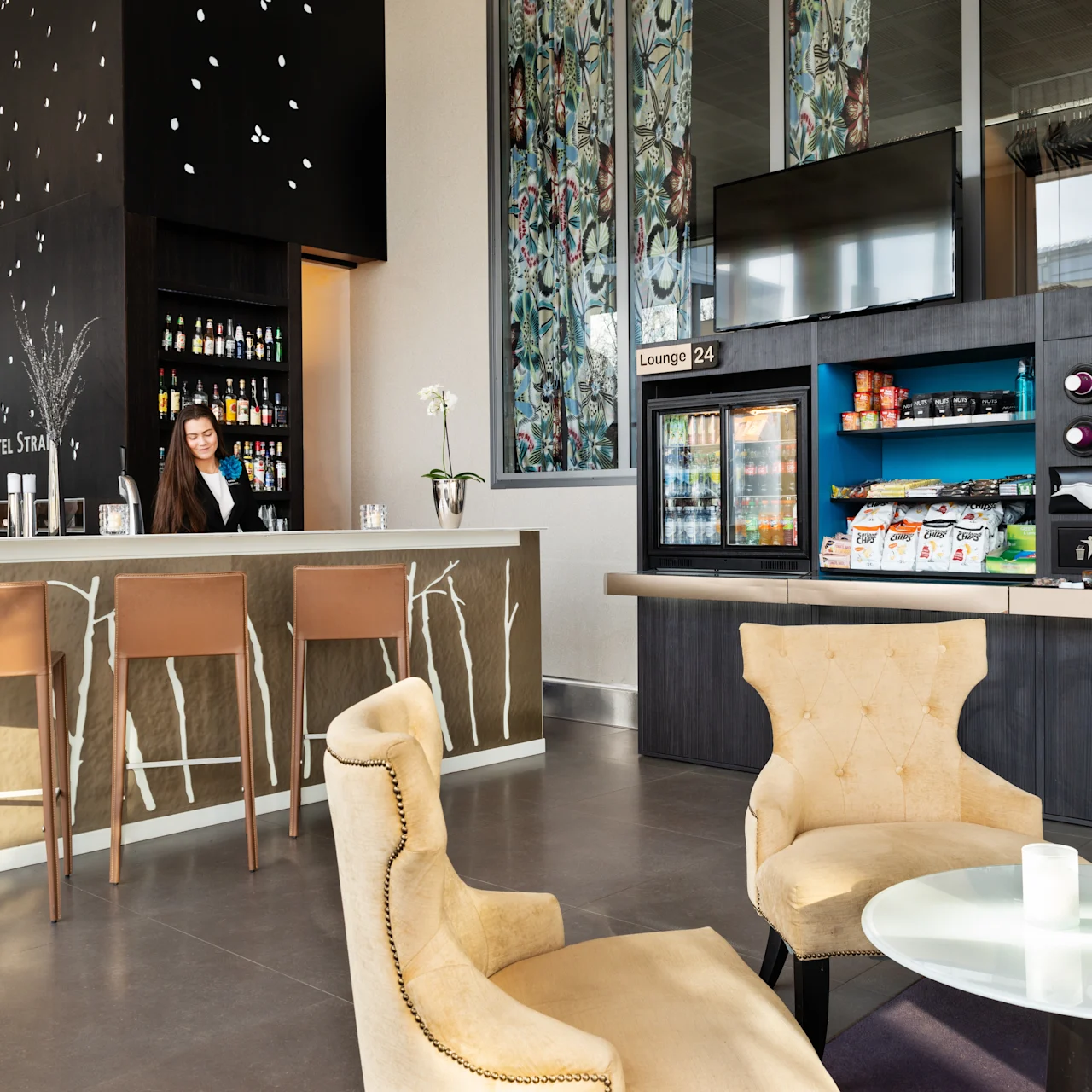 A barista stands behind a hotel bar with shelves of beverages, adjacent to a snack station labeled "Lounge 24." The area features cozy seating with a chair and table. Text: "Quality Hotel Strand."
