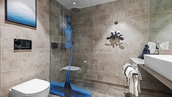 A modern bathroom features a sleek toilet, a glass shower with blue art, and a wide sink with towels. Beige tiles cover the walls and floor.