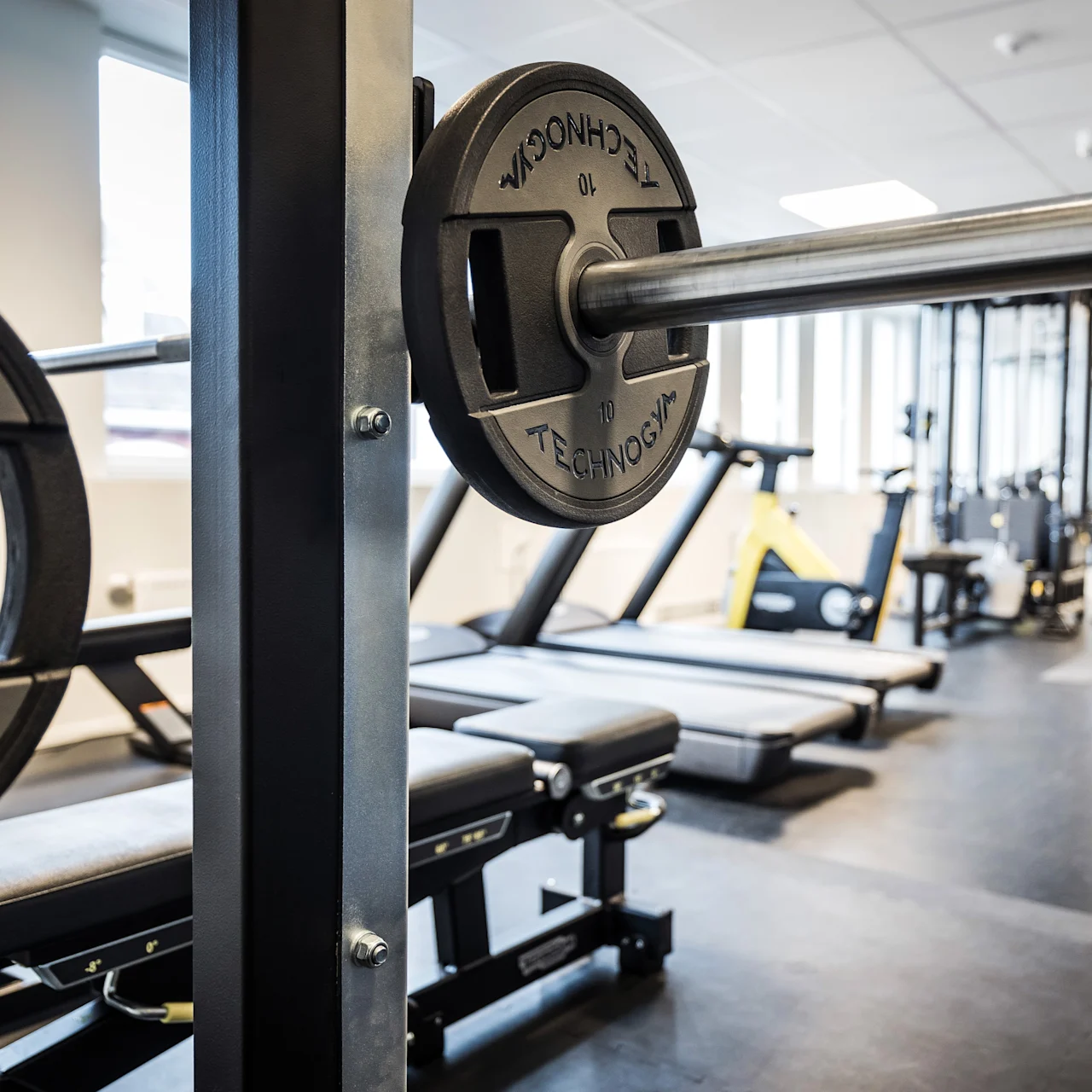 Painonnostotanko, jossa on "Technogym"-painot, lepää telineessä tilavassa kuntosalissa, jossa on juoksumattoja ja kuntoilulaitteita.