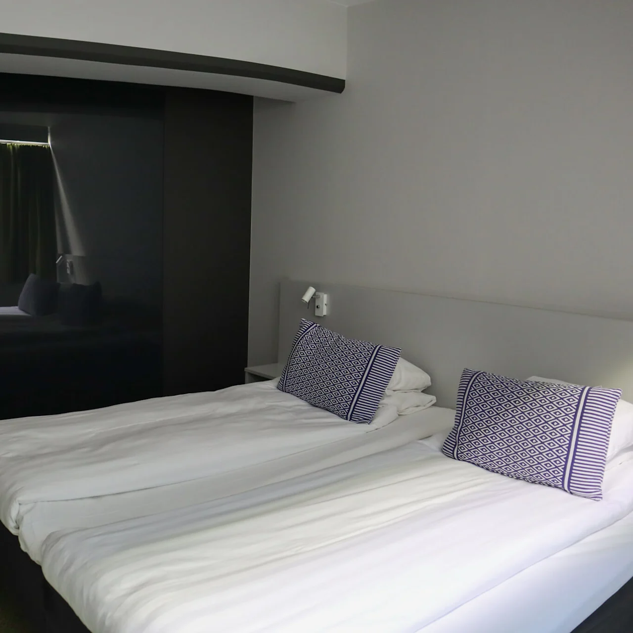 To senger med hvitt sengetøy står i et moderne hotellrom, med mønstrede puter og en svart skyvedør, ved siden av en gang med skogstapet.