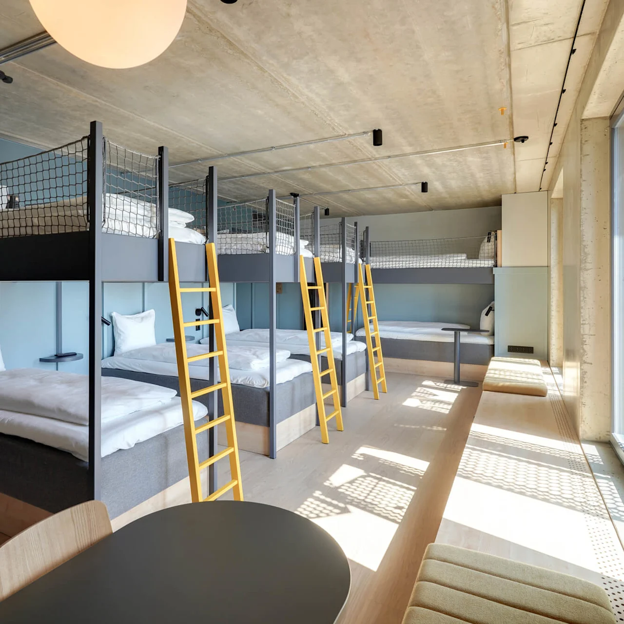 Køjesenge med gule stiger er opstillet langs en væg. Sollys strømmer ind gennem gulv-til-loft-vinduer og oplyser et moderne, minimalistisk rum med trægulve og siddepladser.