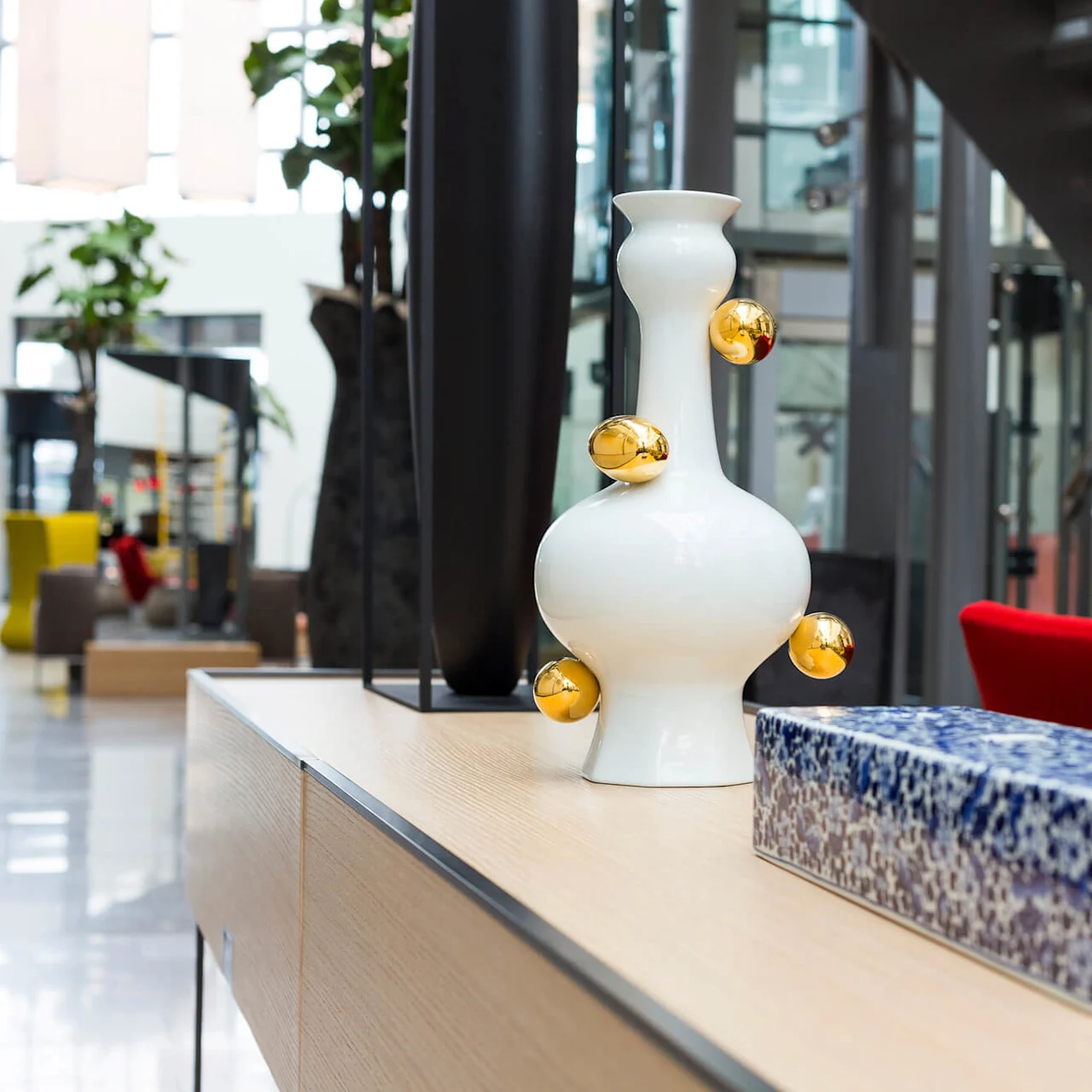 En hvit vase med gullsirkulære aksenter står på et trefat ved siden av en blå mosaikkboks. I bakgrunnen er et moderne lobbyområde med trapper og fargerike sitteplasser.