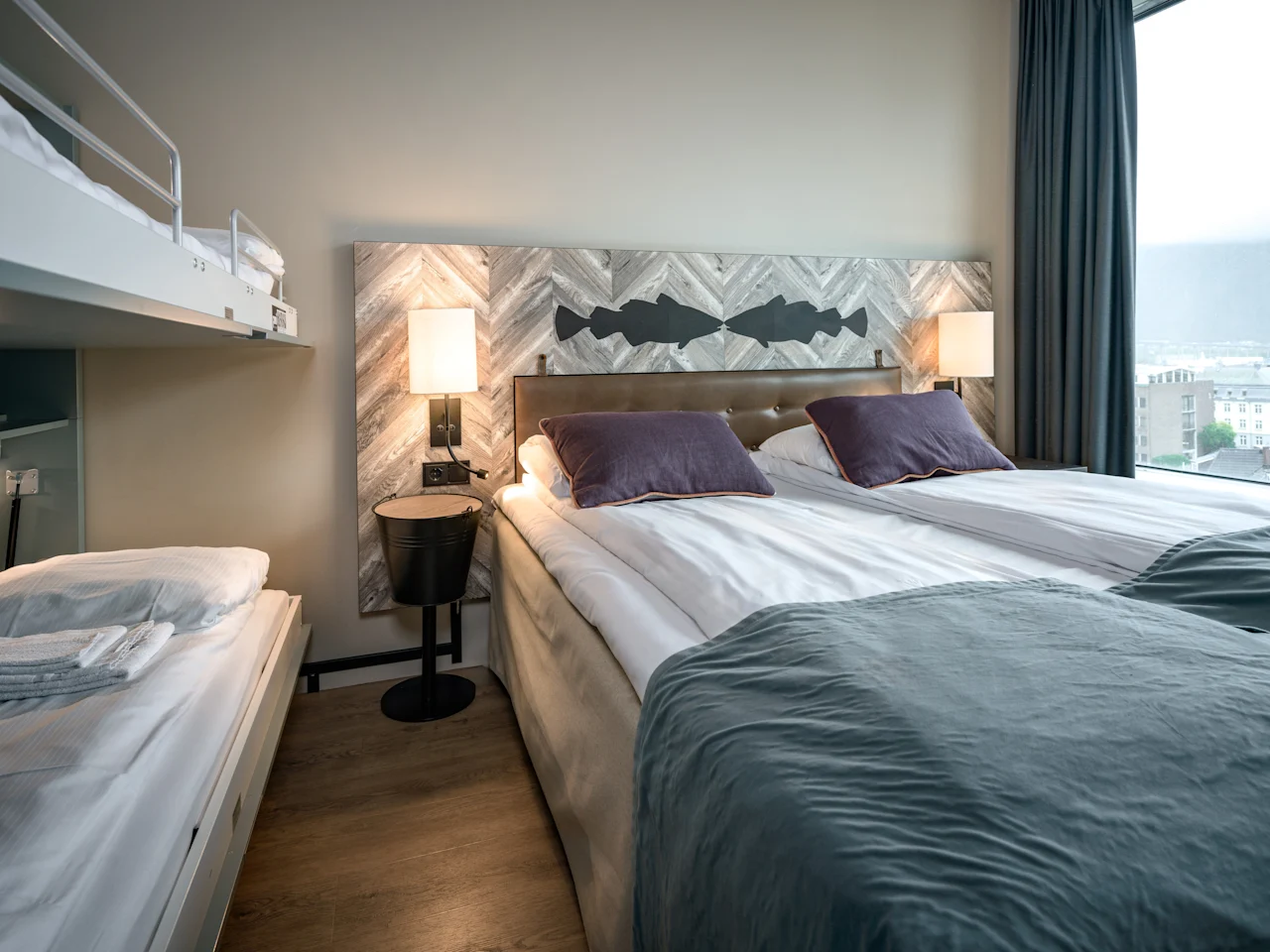 Hotellihuone, jossa on parivuode ja kerrossänky, koristeellinen sängynpääty ja kaupunkinäköala ikkunasta.