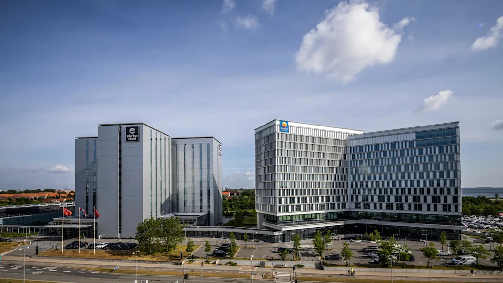 A modern hotel with large glass facades, surrounded by parking spaces and green trees under a blue sky with light clouds.