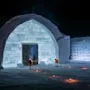 En snebygning med en buet indgang, oplyst af omkringliggende lys på jorden og fakler. Scenen foregår udendørs om natten med snedækket grund og væg.