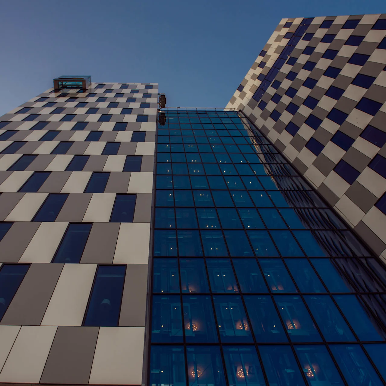 To moderne skyskrabere rejser sig højt, med skiftende lyse og mørke paneler. En glasbro forbinder dem, reflekterende vinduer viser interiørlys, under en klar blå himmel.