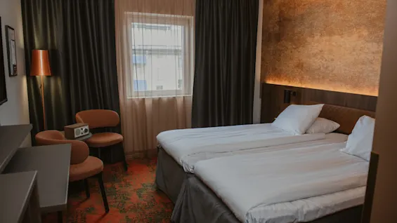 Et komfortabelt hotellrom med to pent oppredde senger, et vindu med både tynne og mørkleggende gardiner, og et lite sitteområde.