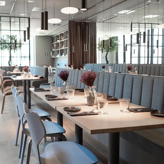 Moderne spiseborde dækket med tallerkener, glas og røde blomsterarrangementer i en elegant restaurant. Stoppede stole står langs bordene. Store vinduer og hængende lys skaber en lys atmosfære.