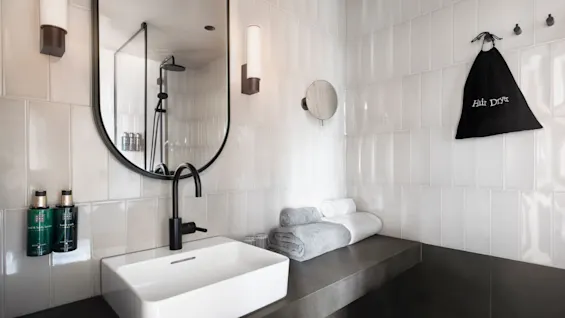 Badrumshandfat med svart kran reflekteras i en väggspegel. Handdukar är prydligt staplade på bänken. Två flaskor märkta "Hand & Body Wash" är monterade på den kaklade väggen. En påse märkt "Hårtork" hänger i närheten.