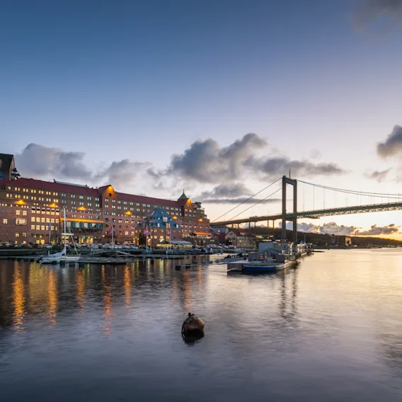 Waterfront hotel building at dusk with a large suspension bridge in the background under a colorful sky.