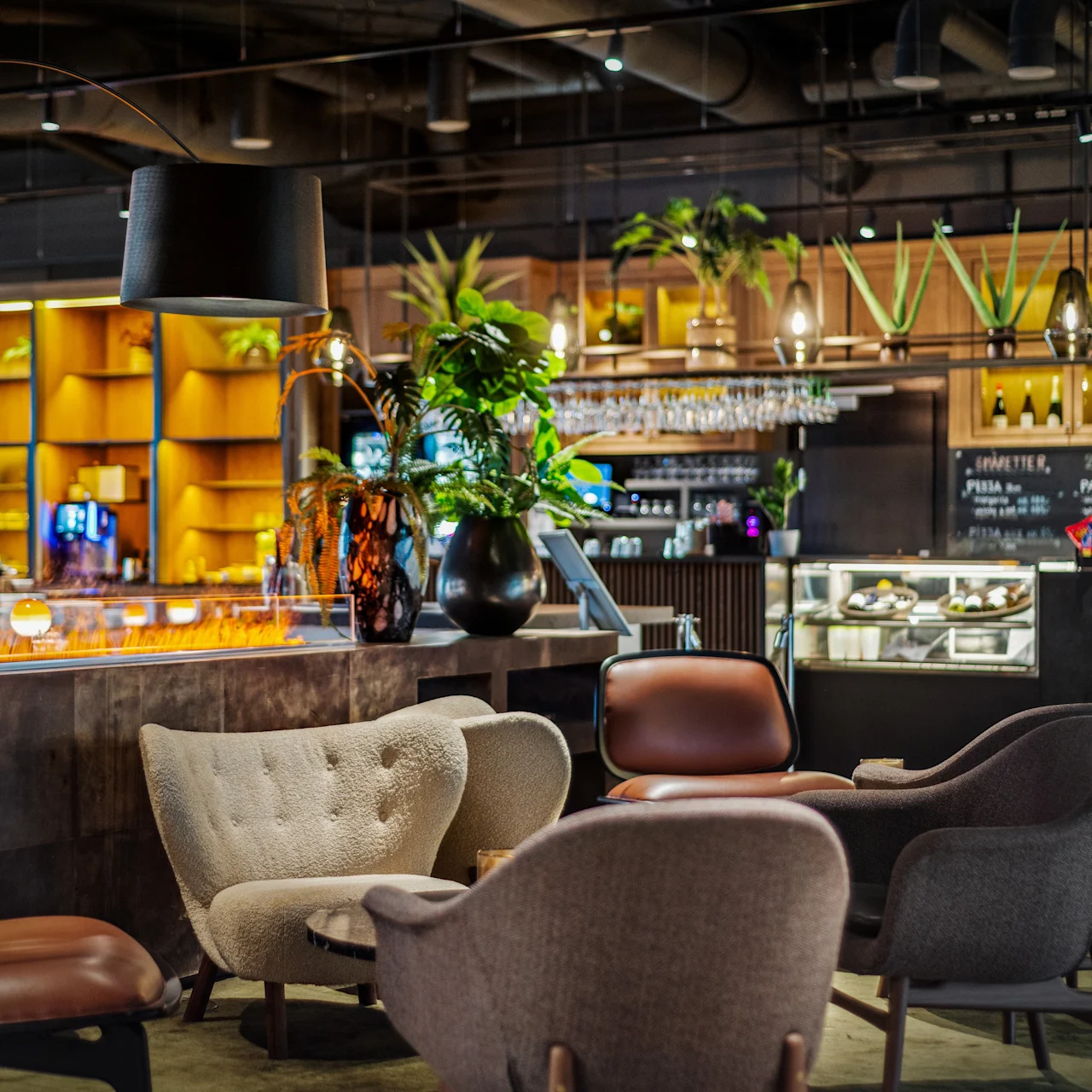 Komfortable stole sidder i en moderne caféindstilling med varm belysning, omgivet af planter og en disk fyldt med drikkevarer og snacks til salg.