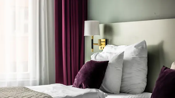 Ett mysigt hotellrum med en säng med vita kuddar och lila kuddfodral. Ljuset från lampan och de mörklila gardinerna mot fönstret ger en varm känsla.