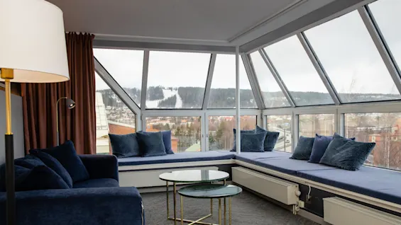 Ett mysigt vardagsrum har en blå soffa och kuddar, med två runda glasbord. Stora fönster avslöjar ett snöigt landskap utanför, vilket ger en lugn atmosfär.