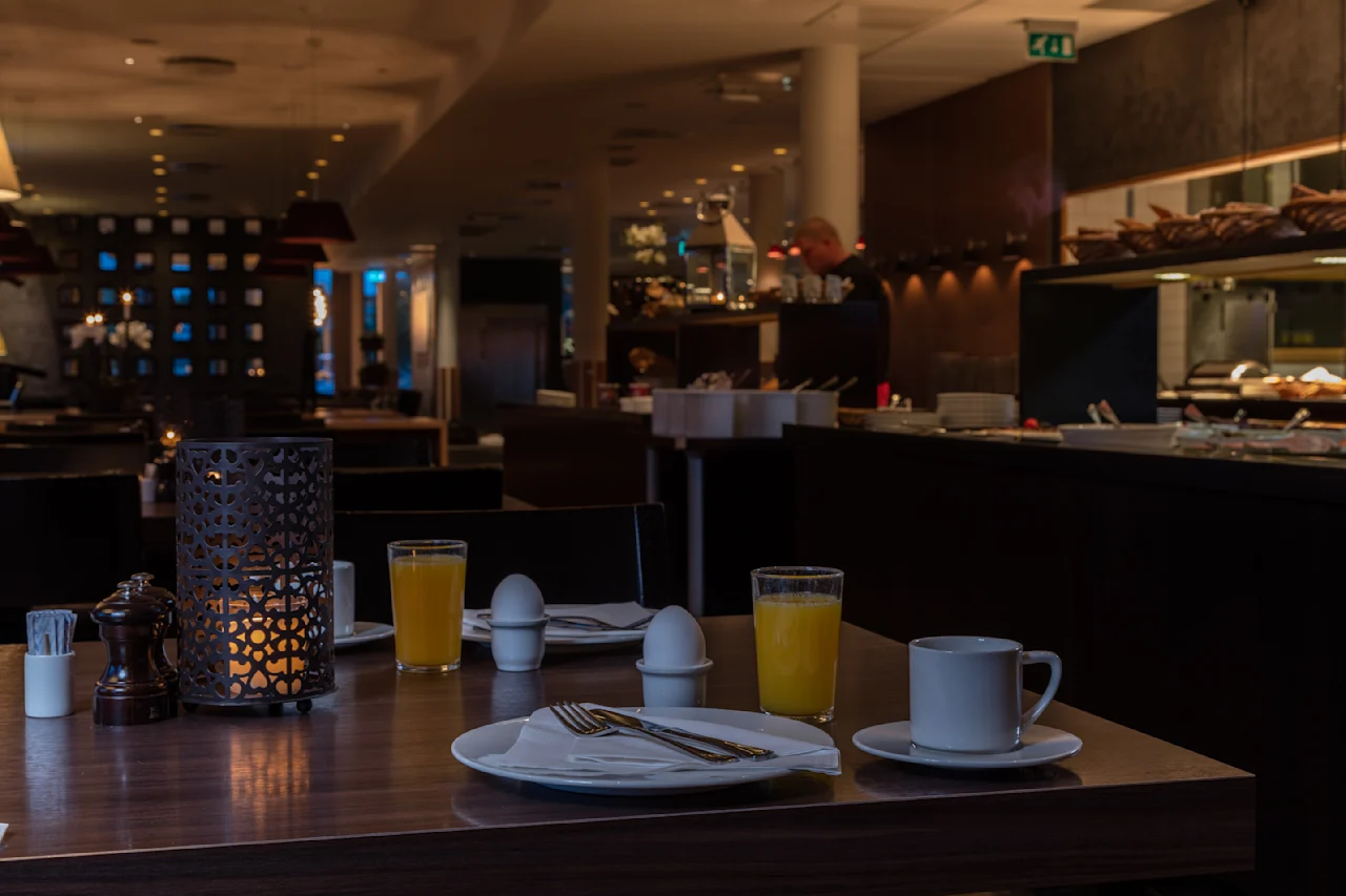 Et bord dekket med bestikk, juice og kaffe med utsikt over en svakt opplyst restaurant. I nærheten har en buffet forskjellige retter. En dekorativ lysestake gir omgivende belysning.