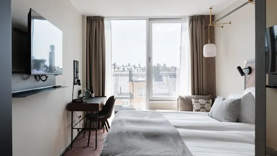 Ett hotellrum med en stor säng, skrivbord och TV, starkt dagsljus från ett fönster som visar stadssilhuett.
