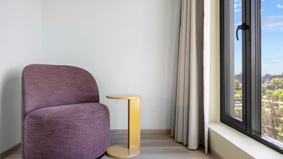 A cozy purple armchair and a golden side table by a large window with a view of a city and trees.