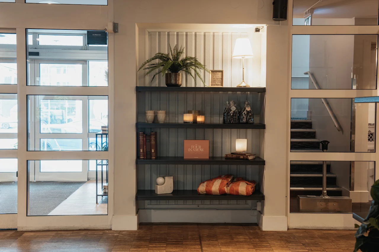 An inviting hotel space with a well-decorated bookshelf, a plant, and a lamp. A book titled FOUR IN A ROW is displayed.