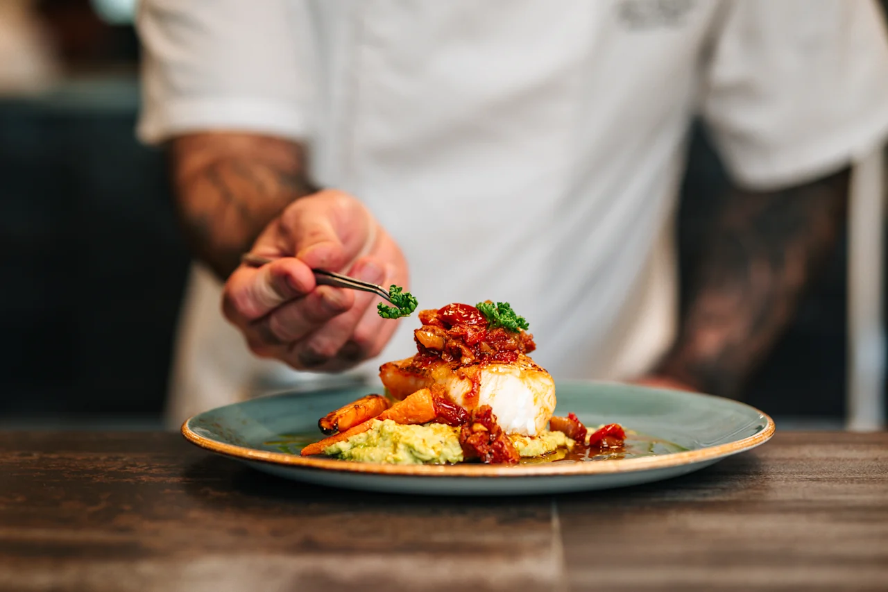 A chef using tweezers to carefully garnish a beautifully plated fish dish.