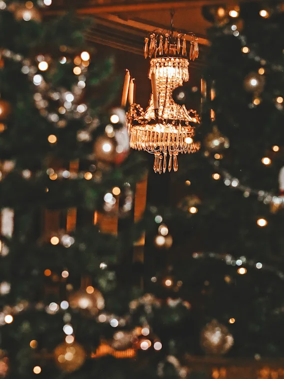 En kristallkrona hänger elegant och avger varmt ljus, omgiven av suddiga, dekorerade julgranar med prydnader som skapar en festlig och lyxig atmosfär.