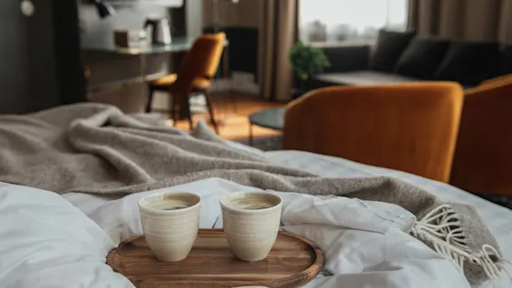 To kopper kaffe på en bakke på en seng på et hotelværelse, hvilket antyder en afslappende morgen eller roomservice.