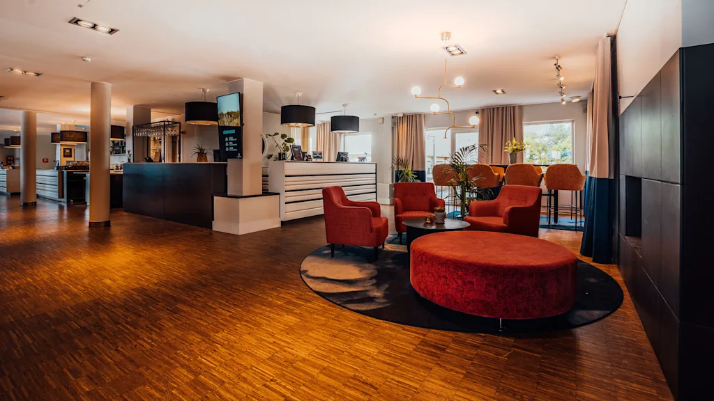Ett modernt lobbyområde med röda fåtöljer, runda bord, trägolv och stämningsbelysning.