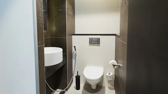 Toilet monteret på en flisebeklædt væg med vægmonteret skylleknap, ved siden af en moderne hvid håndvask og sort børsteholder, i et kompakt og minimalistisk badeværelse.