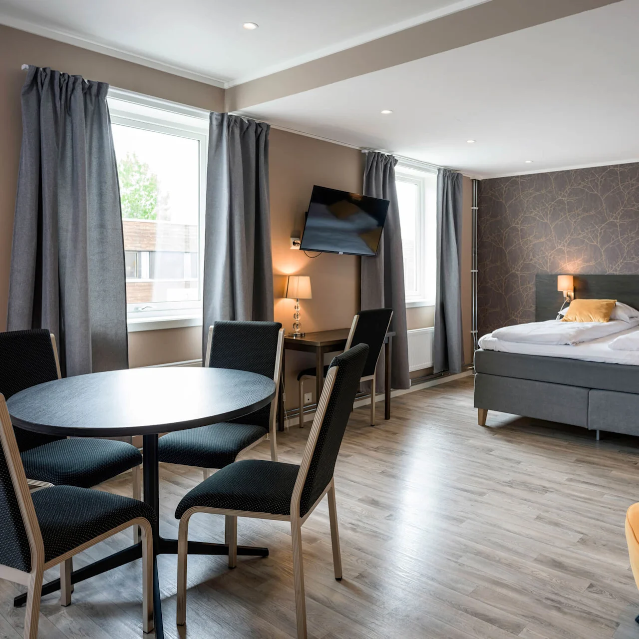 A modern hotel room features a double bed with yellow pillows, a round table with four chairs, a wall-mounted TV, and two large windows with gray curtains.