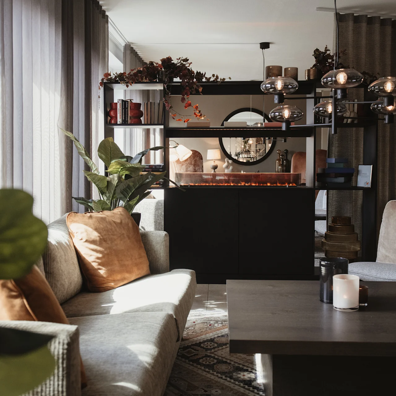 A cozy living room features a gray sofa with brown cushions, plants, and a modern black fireplace in a spacious, sunlit area.
