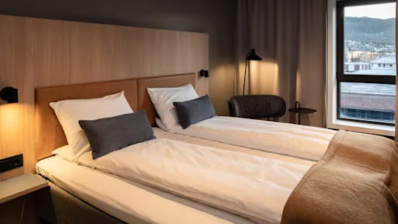 A cozy hotel room with two beds, warm lighting, and a window offering a view of a city and mountains.